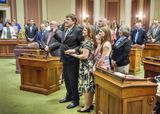 Representative Nelson stands near the front of the House Chamber with one hand on a book and his other hand up. His family stands around him. The House Members also stand at their desks and watch the swearing-in process.